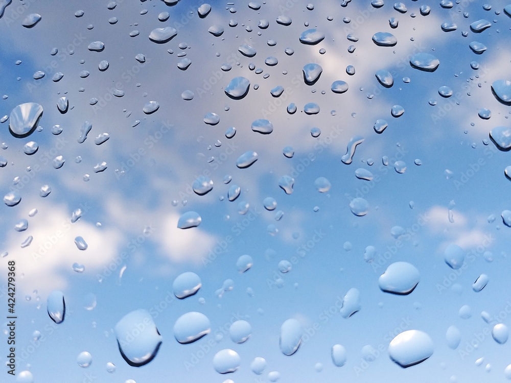 Rain water drops on glass with tropical blue sky background with clouds. Abstract texture of drops of rain on a window glass. Clean background view of water drops on glass over a blue sky.