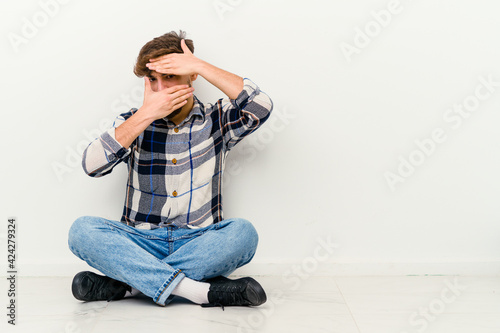 Young Moroccan man sitting on the floor isolated on white background blink at the camera through fingers, embarrassed covering face. © Asier