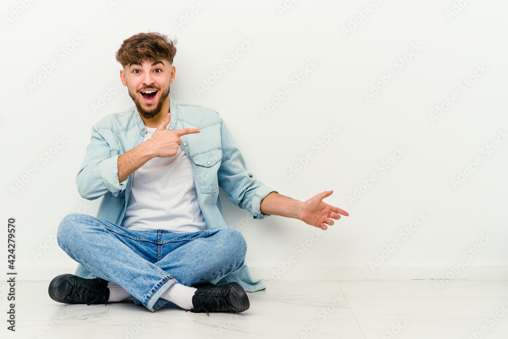 Young Moroccan man sitting on the floor isolated on white background excited pointing with forefingers away. Young Moroccan man sitting on the floor isolated on white background excited pointing with forefingers away.