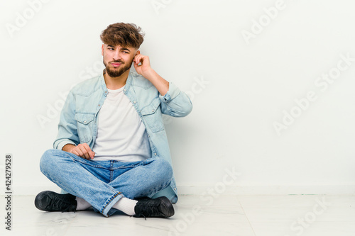 Young Moroccan man sitting on the floor isolated on white background covering ears with fingers, stressed and desperate by a loudly ambient. © Asier