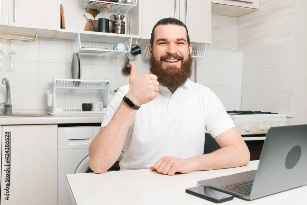 Cheerful young bearded man working from home at laptop and showing thumb up gesture Cheerful young bearded man working from home at laptop and showing thumb up gesture