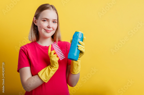 Young blonde happy smiling woman ready to cleaning house with household supplies isolated over yellow color background. Woman Portrait with cleaning brush cleaner in rubber gloves. Copy space