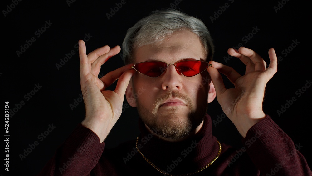 Portrait of seductive young man in trendy stylish clothes wearing sunglasses, charming smile wink blink eye, flirting on black background. Fashion people sincere emotions lifestyle. Attractive guy
