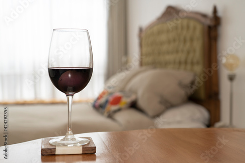 Glass of red wine on a table, behind a bed with a stylish back, and window. Day time. Room place.