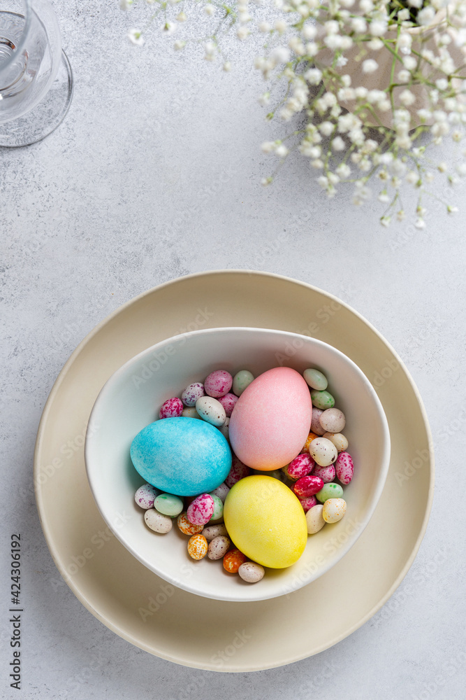 Fototapeta premium Festive Easter table with small white flowers, colored eggs and colorful speckled chocolate candies on ceramic plate. Top view