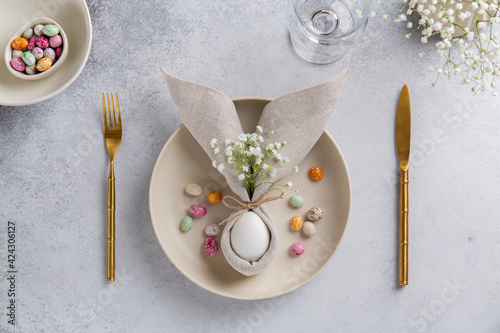 Easter table setting with egg in napkin in the form of an Easter bunny with ears, with small white flowers on ceramic plate and cutlery. Easter table decorations. Top view