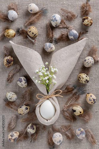 Stylish Easter background with egg in napkin in the form of an Easter bunny with ears, with small white flowers, quail eggs and speckled feathers on sacking. Top view.