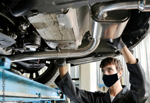 Young Mechanic With Protective Mask for Covid-19 virus on the Mouth Installing Exhaust Pipe Under a Car. New generation of sportive mufflers: double Car Exhaust Pipe chromed made of stainless steel photo