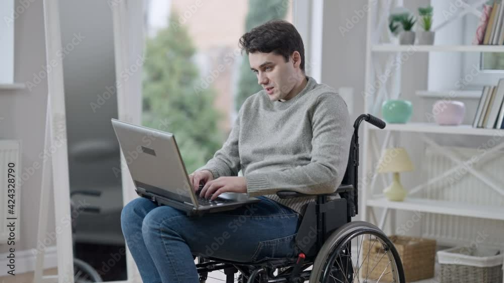 Portrait of positive handsome young paraplegic man in wheelchair talking messaging online using laptop. Brunette Caucasian paralyzed disabled guy indoors at home. Handicap and lifestyle Portrait of positive handsome young paraplegic man in wheelchair talking messaging online using laptop. Brunette Caucasian paralyzed disabled guy indoors at home. Handicap and lifestyle