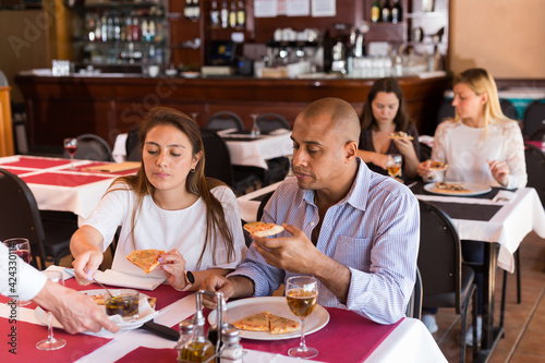 Positive latin american married couple sitting in cozy restaurant and eating pizza with wine