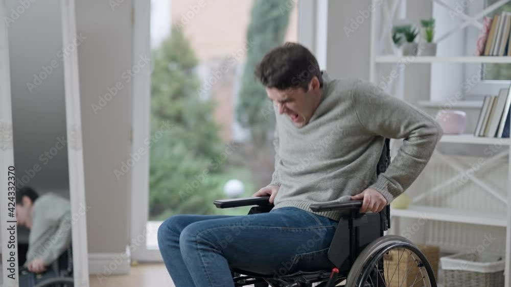 Handsome concentrated young paraplegic man with paralyzed legs trying to stand up of wheelchair indoors. Portrait of confident motivated Caucasian invalid recovering after trauma. Effort and medicine Handsome concentrated young paraplegic man with paralyzed legs trying to stand up of wheelchair indoors. Portrait of confident motivated Caucasian invalid recovering after trauma. Effort and medicine