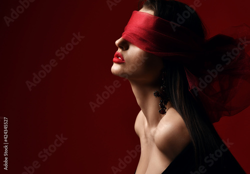 Profile of excited woman with naked shoulders and breast holds and eyes covered with red scarf, blindfold over dark background with copy space. Fashion, vogue, sexy stylish look for woman concept photo