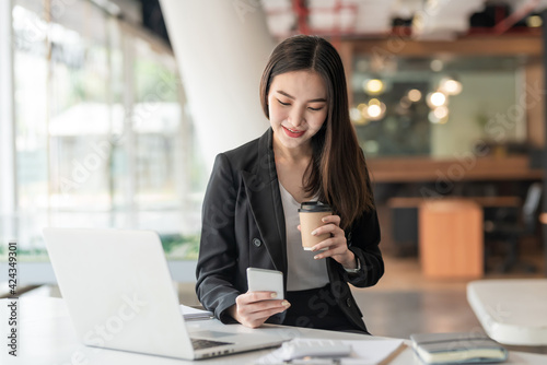 Young Asian businesswoman holding coffee and using smartphone, laptop at office.
