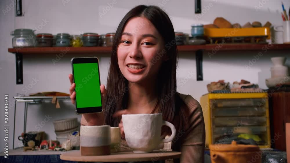 A young female ceramicist is holding smart phone with green screen and new collection of ceramic mugs in the workshop A young female ceramicist is holding smart phone with green screen and new collection of ceramic mugs in the workshop