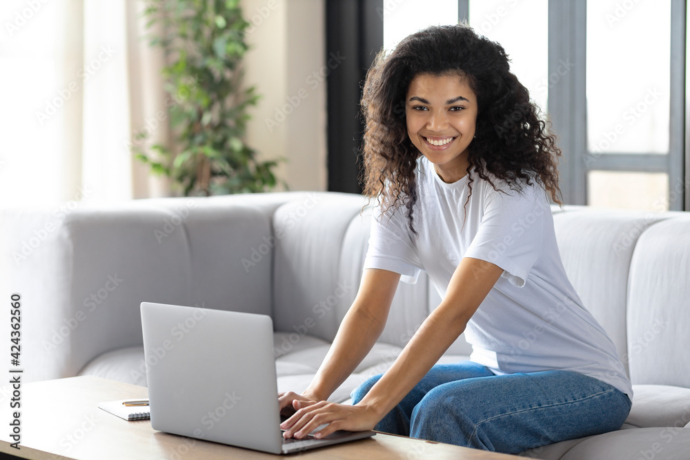 Happy woman using laptop while sitting at home sitting on the couch. Beautiful African American young woman looking at the camera Happy woman using laptop while sitting at home sitting on the couch. Beautiful African American young woman looking at the camera