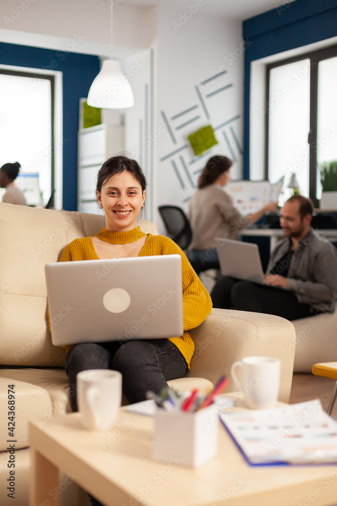 Fototapeta premium Young woman entrepreneur sitting on couch in middle of business start up office looking at camera smiling holding laptop. Multiethnic coworkers talking about startup financial company in modern office