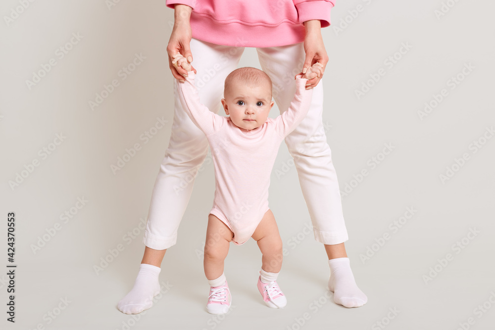 Mom teaches her daughter to walk, faceless female wearing white pants, holding her baby's hands and walking indoor against white wall, kid looks at camera and likes to go. Mom teaches her daughter to walk, faceless female wearing white pants, holding her baby's hands and walking indoor against white wall, kid looks at camera and likes to go.