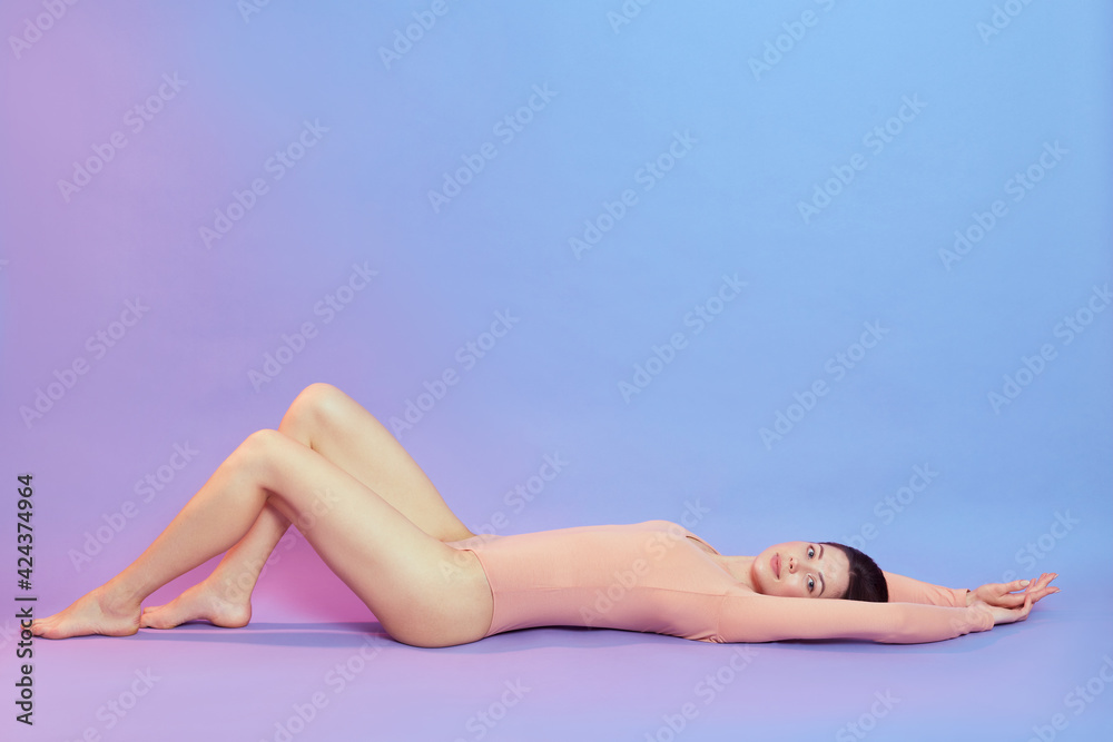 Obraz premium Beautiful young woman with long legs wearing beige bodysuit lying on floor and looking up, female with perfect body raising legs, posing isolated over blue background with pink neon light.