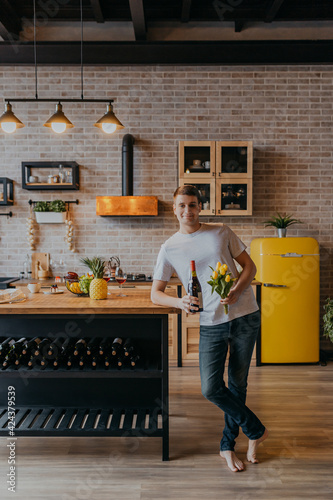 Handsome man with wine bottle and flowers in kitchen. Women's Day card with man with presents in modern kitchen.