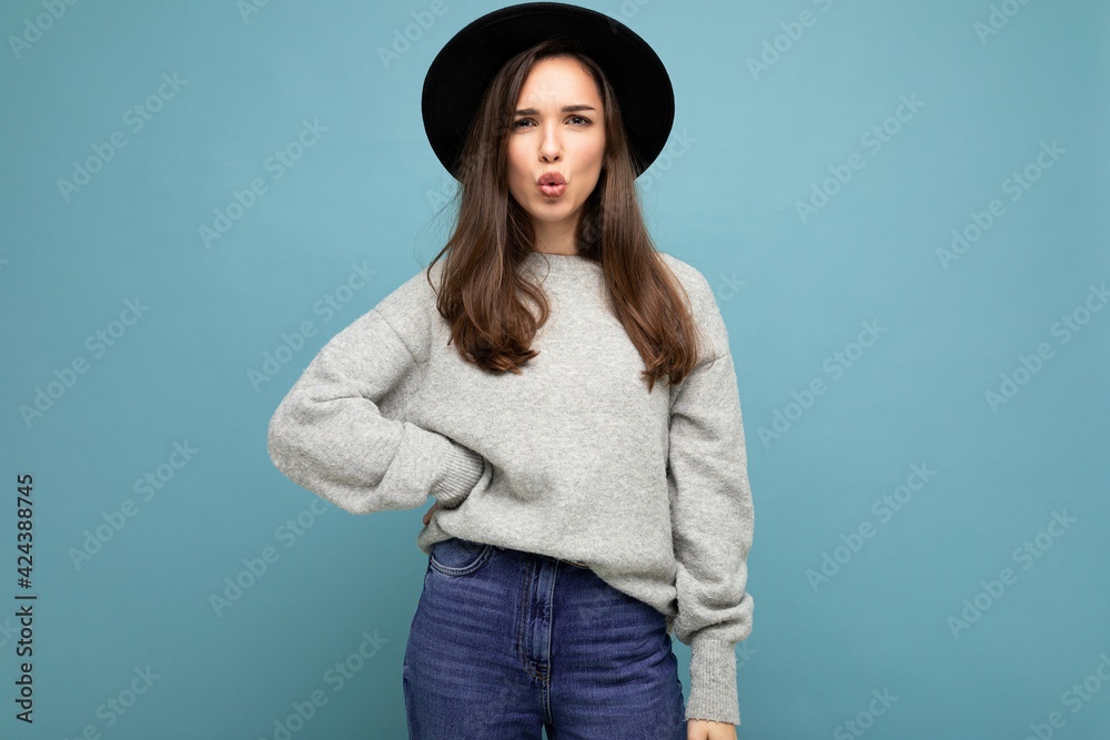 Young fascinating brunet woman. Trendy lady in casual grey sweater and stylish black hat. Female shows facial sincere emotions isolated on blue background with free space and giving kiss Young fascinating brunet woman. Trendy lady in casual grey sweater and stylish black hat. Female shows facial sincere emotions isolated on blue background with free space and giving kiss