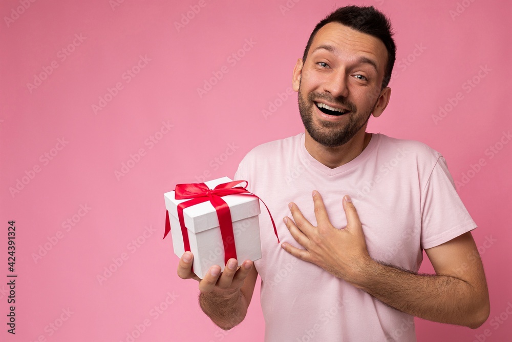 Shot of handsome positive joyful delightful brunette adult unshaven man with beard isolated over pink background wall wearing pink t-shirt holding white gift box with red ribbon and looking at camera