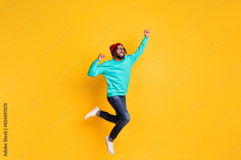 Full size profile photo of brunette optimistic guy jump wear cap spectacles pullover jeans sneakers isolated on yellow color background