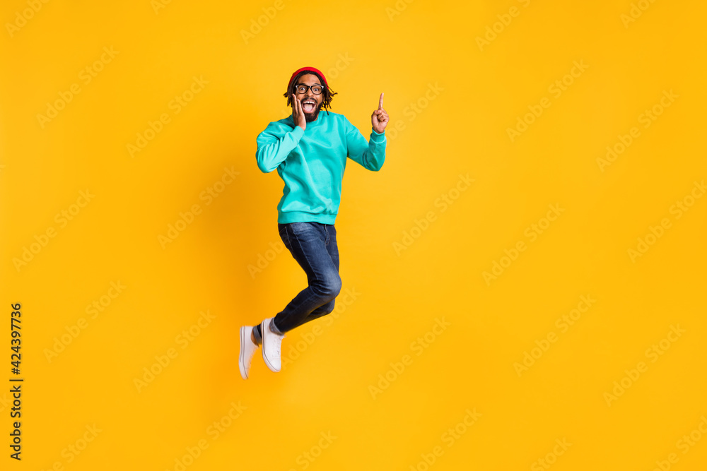 Full size profile photo of brunette optimistic guy jump point empty space wear cap spectacles pullover jeans isolated on yellow background