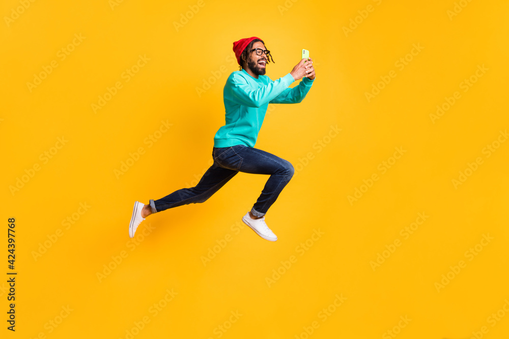 Full size profile photo of brunette optimistic guy jump run take photo wear cap spectacles pullover jeans isolated on yellow background