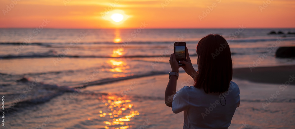 Silhouette of happy young woman taking photo by smartphone.Tourist enjoy beautiful sunset at the beach. Travel, relaxing, vacation summer concept