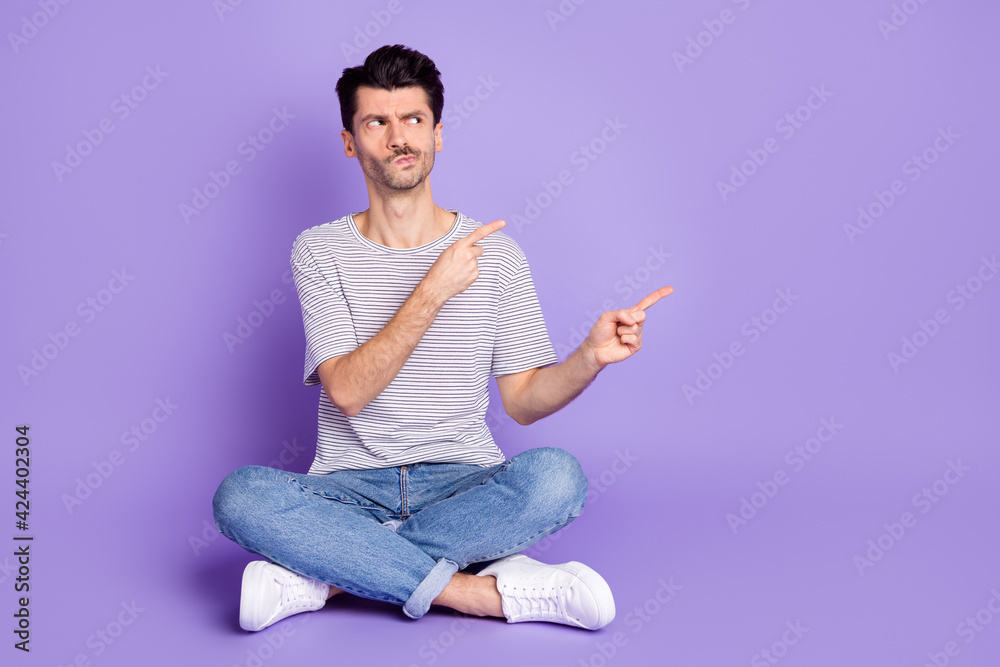 Photo of guy sit floor indicate fingers look empty space wear striped t-shirt jeans footwear isolated violet background Photo of guy sit floor indicate fingers look empty space wear striped t-shirt jeans footwear isolated violet background