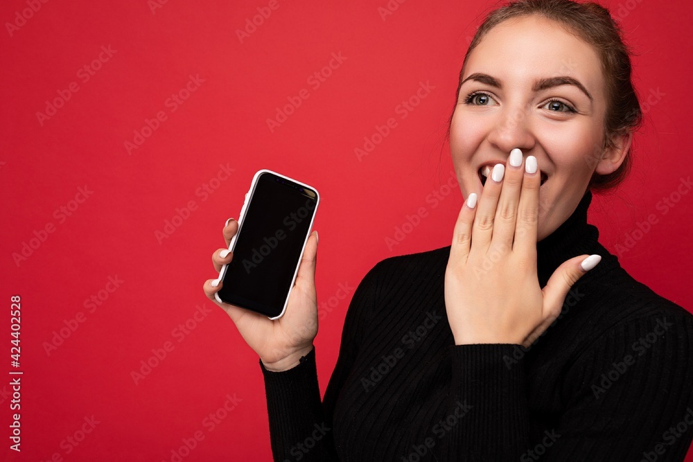 Photo of amazed attractive positive young brunette woman wearing black sweater standing isolated over red background showing mobile phone with empty screen for mockup Photo of amazed attractive positive young brunette woman wearing black sweater standing isolated over red background showing mobile phone with empty screen for mockup