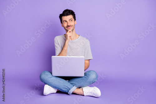Photo of guy sit floor hand chin hold pc look up empty space wear white t-shirt jeans sneakers isolated purple background