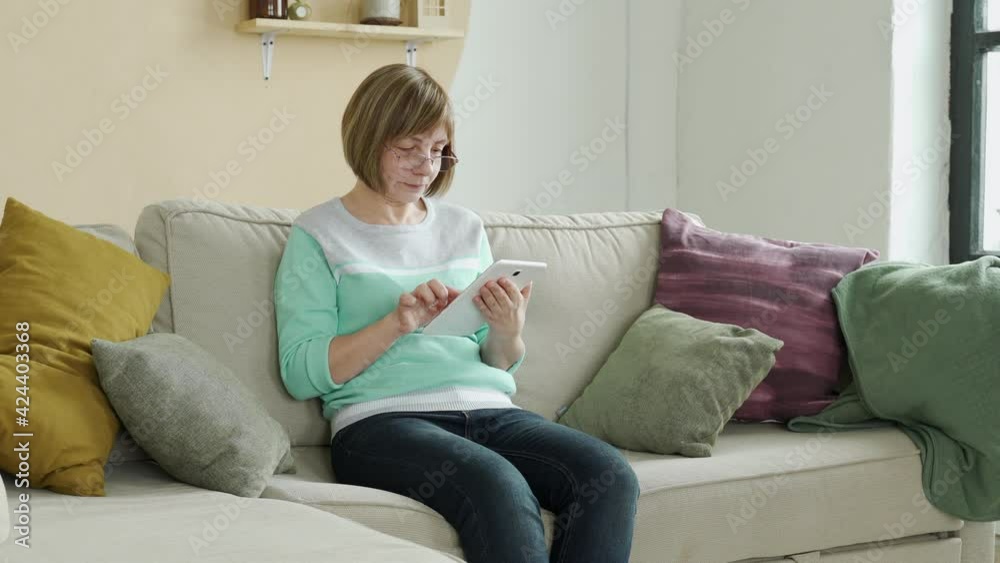 Smiling elderly woman wearing glasses using digital tablet at home sitting on the couch Smiling elderly woman wearing glasses using digital tablet at home sitting on the couch