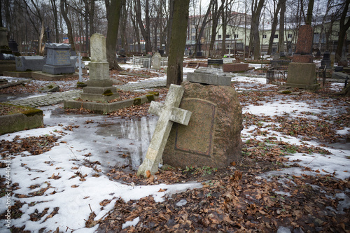Broken stone cross leaning against gravestone in cemetery covered with snow and dry leaves - Smolenskoe Lutheran Cemetery, Russia, Saint Petersburg, March 2021