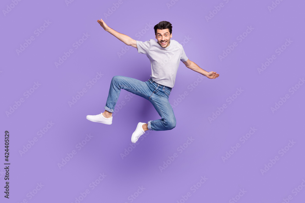 Photo of funky crazy guy jump raise hands plane pose wear striped t-shirt jeans sneakers isolated purple background