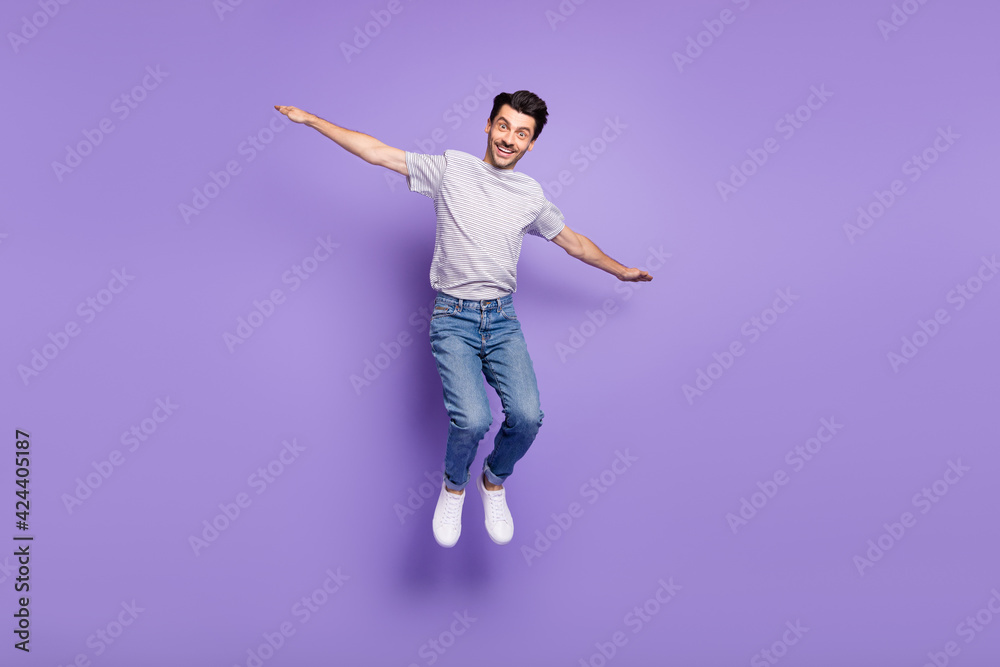Photo of cheerful positive guy jump airplane pose wear t-shirt denim jeans sneakers isolated purple background Photo of cheerful positive guy jump airplane pose wear t-shirt denim jeans sneakers isolated purple background