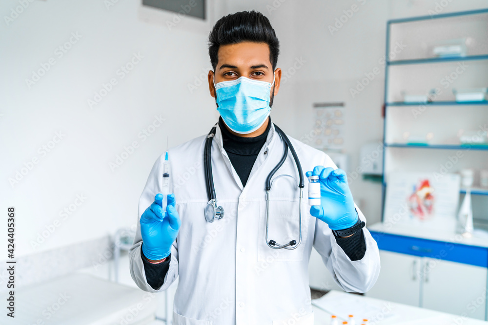 Portrait of a doctor in medical clothes and in a protective mask, holding a vaccine and an injection in his hands. Medicine, health, modern hospital Portrait of a doctor in medical clothes and in a protective mask, holding a vaccine and an injection in his hands. Medicine, health, modern hospital