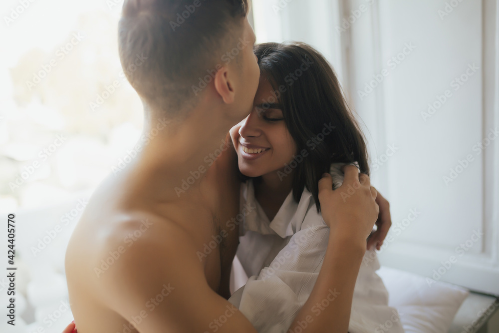 Happy couple of lovers is sitting on the windowsill and hugging. Woman with closed eyes is smiling while her man is kissing her on the forehead. Happy couple of lovers is sitting on the windowsill and hugging. Woman with closed eyes is smiling while her man is kissing her on the forehead.