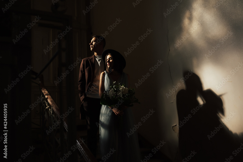 Beautiful and elegant bride in a long blue dress and hat with bouquet in hands and her handsome groom in a suit are standing in the dark room with sunlight.