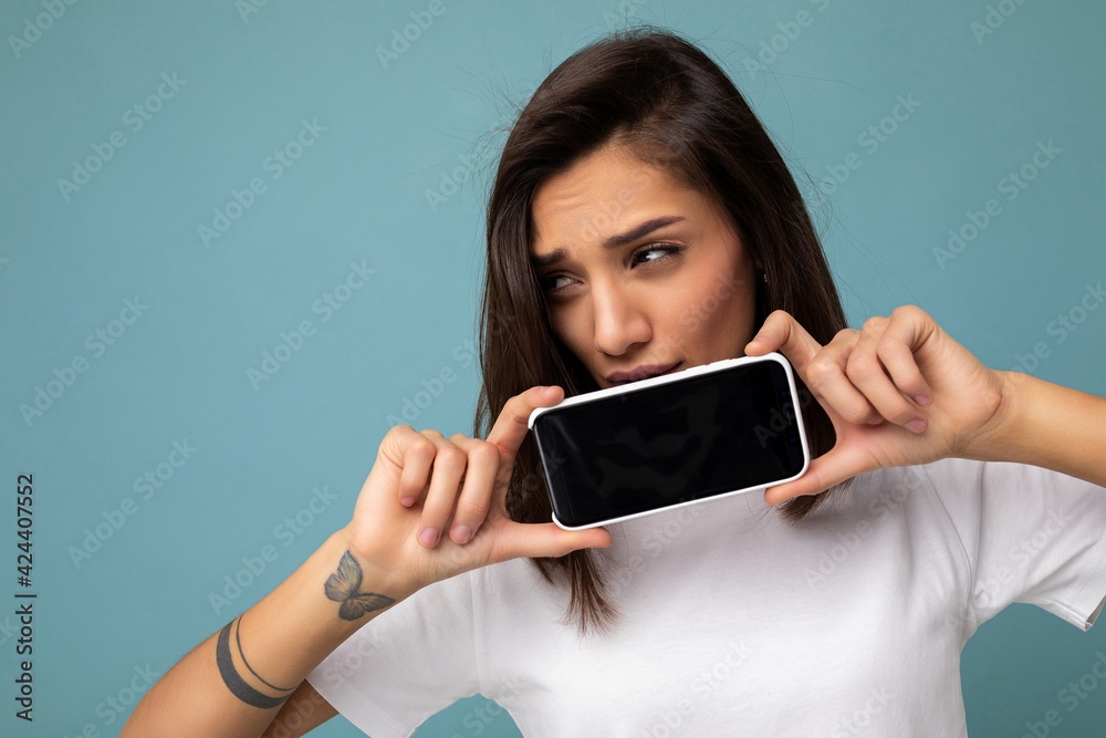portrait Photo of sad pretty young brunette woman good looking wearing white t-shirt standing isolated on blue background with copy space holding smartphone showing phone in hand with empty screen portrait Photo of sad pretty young brunette woman good looking wearing white t-shirt standing isolated on blue background with copy space holding smartphone showing phone in hand with empty screen