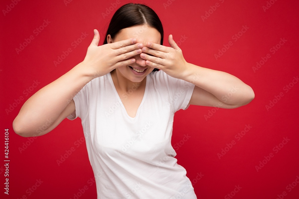 Photo of positive smiling young beautiful brunette woman with sincere emotions wearing white t-shirt for mockup isolated over red background with empty space and covering eyes Photo of positive smiling young beautiful brunette woman with sincere emotions wearing white t-shirt for mockup isolated over red background with empty space and covering eyes