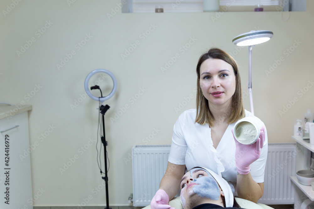 Facial skin care. Beautiful woman with cosmetic facial mask in the spa salon. Woman having a facial cosmetic mask. A beautician in a white medical gown makes a cosmetic procedure in a beauty salon. Facial skin care. Beautiful woman with cosmetic facial mask in the spa salon. Woman having a facial cosmetic mask. A beautician in a white medical gown makes a cosmetic procedure in a beauty salon.