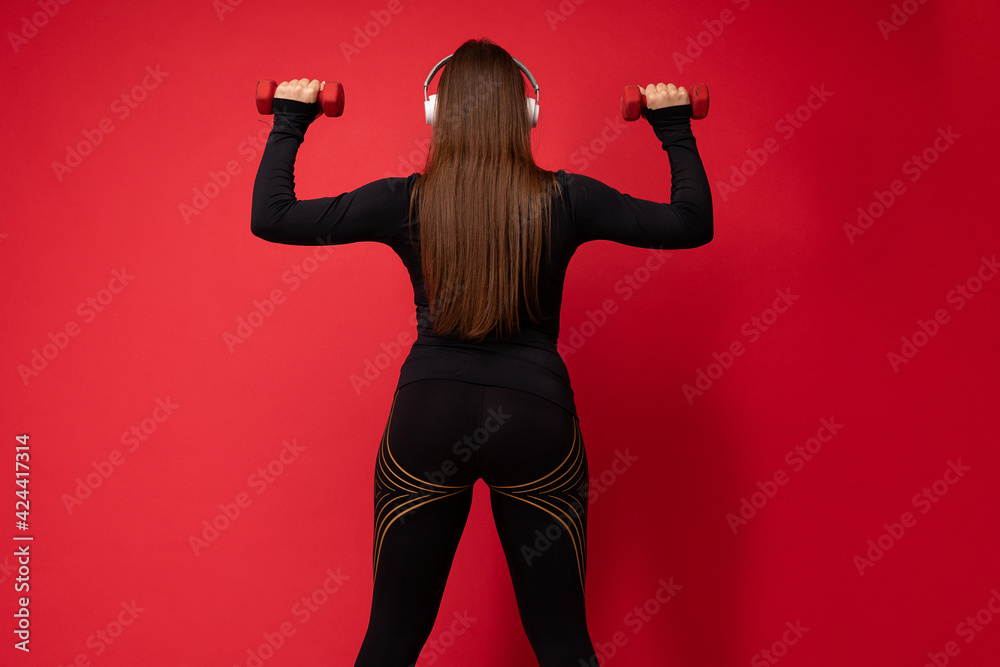 Beautiful happy smiling young brunet woman wearing black sport clothes isolated on red background wall doing fitness using dumbbells wearing white bluetooth headsets listening to music Beautiful happy smiling young brunet woman wearing black sport clothes isolated on red background wall doing fitness using dumbbells wearing white bluetooth headsets listening to music