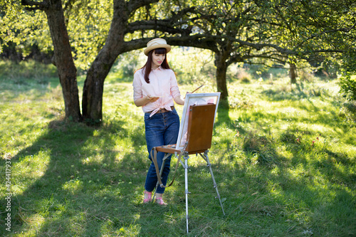 Full-length horizontal shot of pretty young caucasian woman, wearing straw hat, denim jeans and pink shirt, painting on canvas outside in the park.