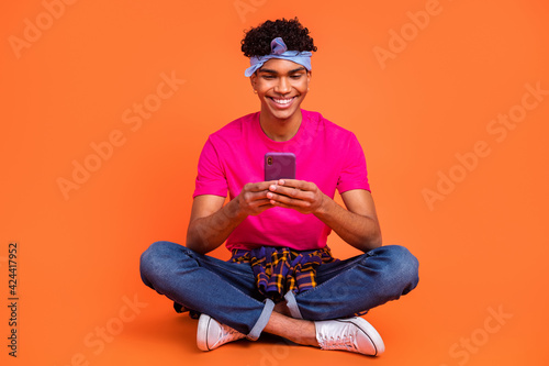 Full length body size photo of young hipster browsing internet with smartphone smiling isolated bright orange color background
