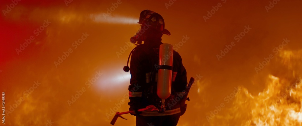Obraz premium Dramatic silhouette of American firefighter in full gear exploring the huge fire zone