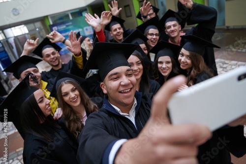 group of happy international students in mortar boards and bachelor gowns with diplomas taking selfie by smartphone