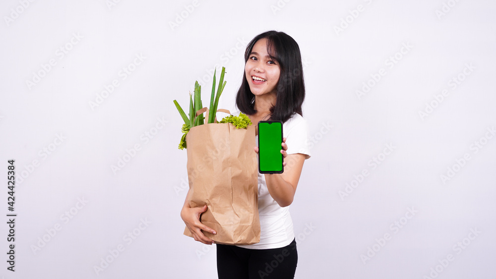 Asian woman with paper bag of fresh vegetables and holding a green screen phone with isolated white background Asian woman with paper bag of fresh vegetables and holding a green screen phone with isolated white background