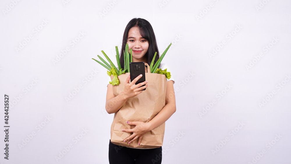 Asian woman with paper bag of fresh vegetables and holding a phone with isolated white background Asian woman with paper bag of fresh vegetables and holding a phone with isolated white background