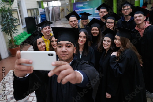 group of happy international students in mortar boards and bachelor gowns with diplomas taking selfie by smartphone